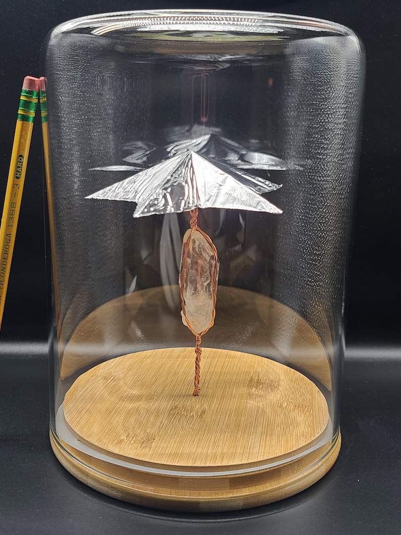 May include: A clear glass cloche with a wooden base houses a quartz crystal wrapped in copper wire. A silver foil structure is suspended above the crystal. Two pencils are visible to the left of the cloche.