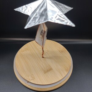 May include: A unique decorative object featuring a silver, star-shaped element atop a clear crystal, supported by a copper wire. The crystal and wire are mounted on a round, light-colored wooden base. The background is black.
