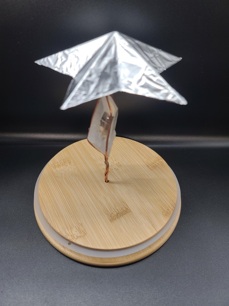 May include: A unique decorative object featuring a silver, star-shaped element atop a clear crystal, supported by a copper wire. The crystal and wire are mounted on a round, light-colored wooden base. The background is black.