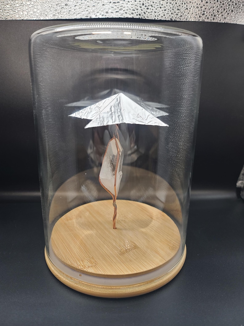 May include: A clear glass cloche with a wooden base houses a quartz crystal suspended by copper wire. A silver foil structure is positioned above the crystal. The cloche sits on a dark surface.