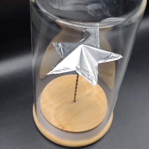 May include: A clear glass cloche with a wooden base and a silver star sculpture inside. The star is attached to a thin, twisted copper wire. The cloche sits on a dark surface.