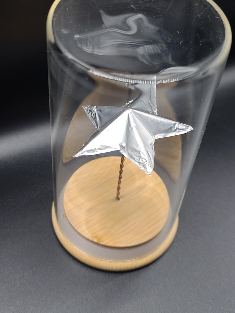 May include: A clear glass cloche with a wooden base and a silver star sculpture inside. The star is attached to a thin, twisted copper wire. The cloche sits on a dark surface.