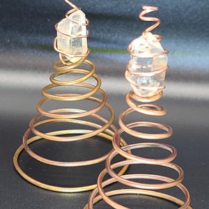 May include: Two handcrafted copper spiral sculptures, each featuring a clear quartz crystal at the top. The crystals are wrapped in copper wire, and the spirals are made of copper. The sculptures are displayed against a dark background.