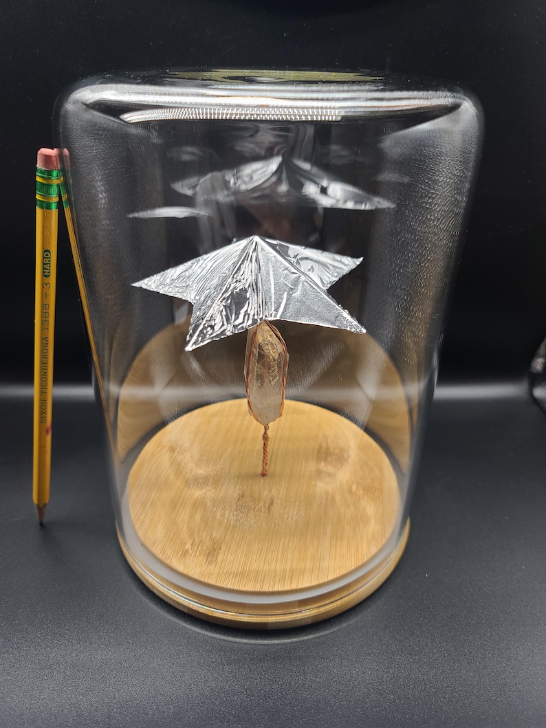 May include: A clear glass cloche with a wooden base houses a crystal suspended by copper wire. A small, silver foil structure sits above the crystal. A yellow pencil with black text is next to the cloche. The cloche is on a dark surface.