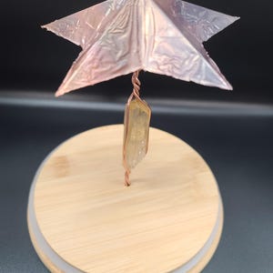 May include: A handcrafted decorative piece featuring a copper wire stem, a clear quartz crystal, and a metallic pink star-shaped top. The piece is mounted on a round wooden base with a white trim, creating a unique artistic display.