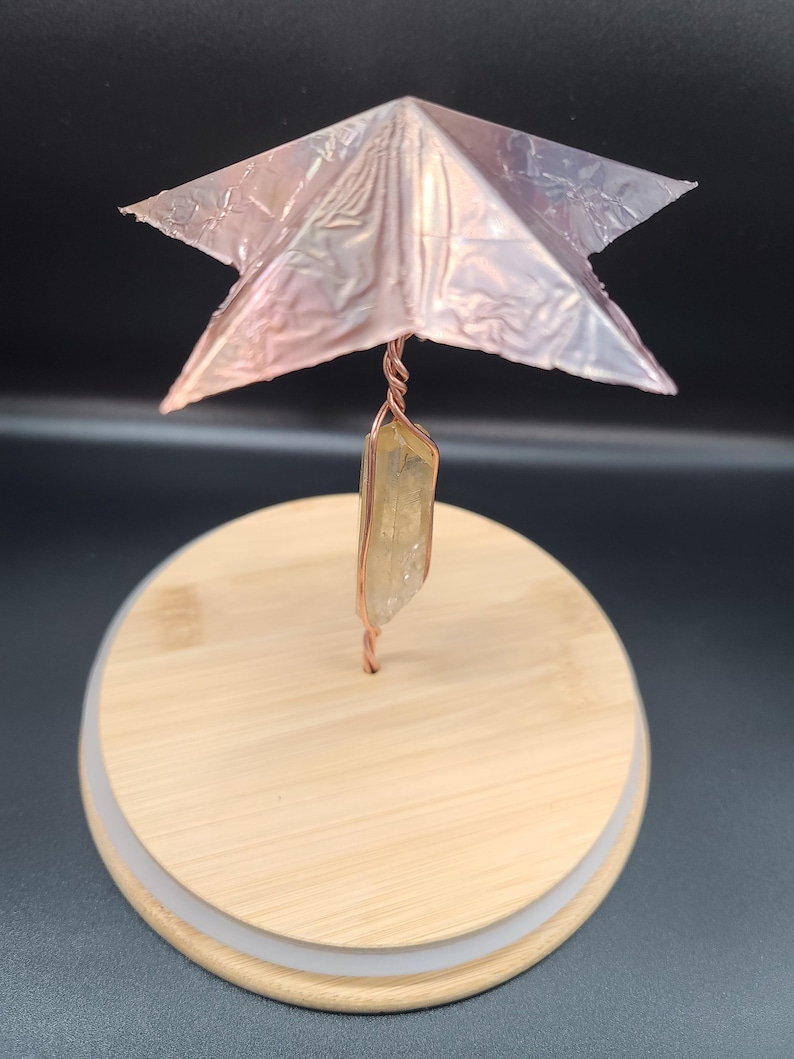 May include: A handcrafted decorative piece featuring a copper wire stem, a clear quartz crystal, and a metallic pink star-shaped top. The piece is mounted on a round wooden base with a white trim, creating a unique artistic display.