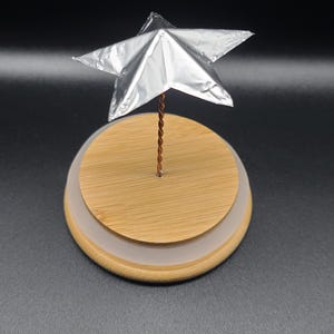May include: A silver star ornament on a wooden base with a clear, frosted ring. The star is made of a reflective material and is attached to the base with a twisted copper wire. The base is round and has a natural wood grain.