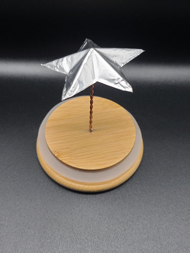 May include: A silver star ornament on a wooden base with a clear, frosted ring. The star is made of a reflective material and is attached to the base with a twisted copper wire. The base is round and has a natural wood grain.
