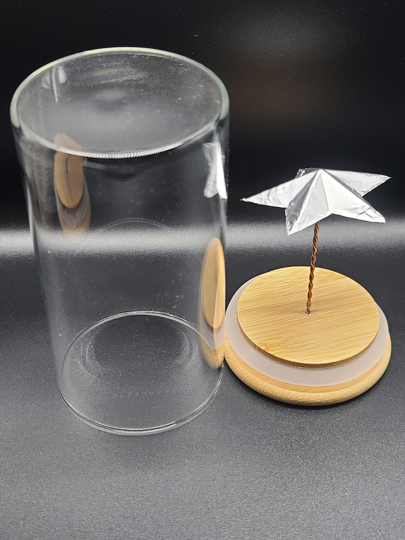 May include: A clear glass terrarium with a wooden base and a small silver origami star on a wire stem. The terrarium is cylindrical and open at the top. The wooden base has a light brown top and a white ring.