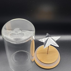 May include: A clear glass cloche with a wooden base and a silver star decoration. The cloche is open, revealing the wooden base with a silver star attached to a thin wire. The star is made of folded silver foil.