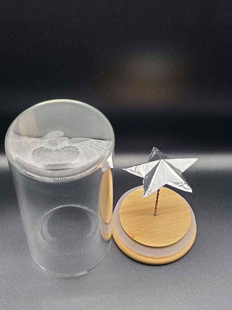 May include: A clear glass cloche with a wooden base and a silver star decoration. The cloche is open, revealing the wooden base with a silver star attached to a thin wire. The star is made of folded silver foil.