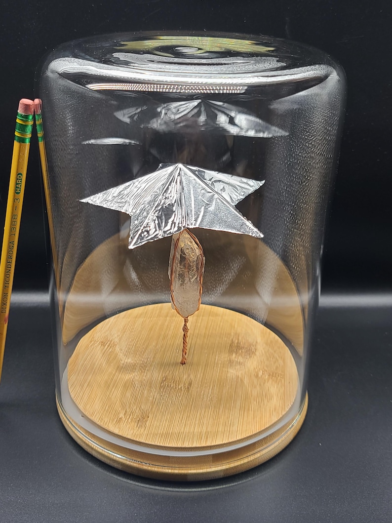 May include: A clear glass cloche with a wooden base houses a crystal suspended by a copper wire. A silver foil umbrella is positioned above the crystal. A yellow pencil with a pink eraser rests on the side.