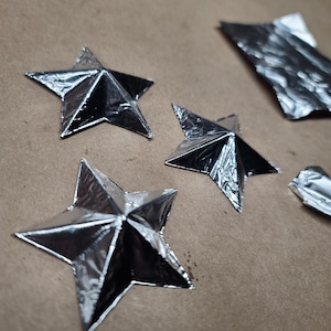 May include: Close-up of several silver foil stars on a brown surface. The stars are in various sizes and shapes, some folded and others flat. One white star is also visible. The stars are likely for decoration or crafts.