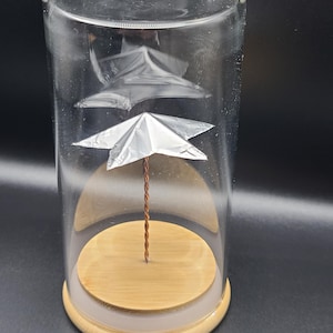 Può includere: Un cilindro di vetro trasparente con una base in legno ospita una scultura in miniatura. La scultura presenta due oggetti argentati a forma di stella sospesi all'interno del vetro, collegati da un sottile filo di rame ritorto. La base è di colore marrone chiaro.