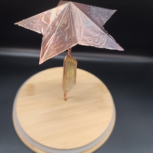 May include: A decorative star sculpture with a copper-colored, metallic finish. The star is mounted on a copper wire that is attached to a light-colored, rectangular crystal. The crystal is attached to a round, wooden base.