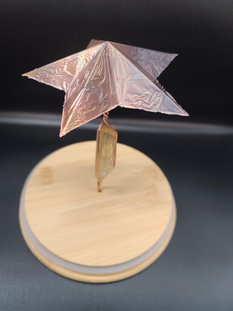 May include: A decorative star sculpture with a copper-colored, metallic finish. The star is mounted on a copper wire that is attached to a light-colored, rectangular crystal. The crystal is attached to a round, wooden base.
