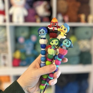 May include: A collection of colorful beaded character wands featuring characters from the animated movie Inside Out. Each wand has a different character figurine, with a variety of colors including blue, red, yellow, green, and pink. The wands are held in a hand.