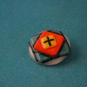 May include: A glass button with a geometric design. The button is white with black lines and a red and yellow square in the center. The square has a black plus sign in the center.