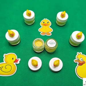 May include: A set of seven yellow rubber ducky bath toys, six in small jars and one large ducky on a green background. The jars have white lids with a small yellow ducky on top. The large ducky has a red beak and black eyes. The text on the bottom of the image reads "©2024 Nyx DarkSkye of Nyx's Notions™".