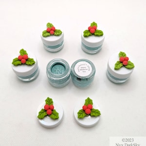 May include: Several small, clear jars filled with a teal-colored substance, each topped with a white lid decorated with a green holly leaf and red berry design. One jar is open, revealing the contents. The text on the label reads "Merry Mistletoe".