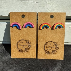 May include: Two pairs of rainbow-colored wooden earrings. The first pair is pink, purple, blue, and green. The second pair is red, orange, yellow, green, blue, and purple. Both pairs are displayed on brown cards with the text "S.M Designs and decor" printed on them.