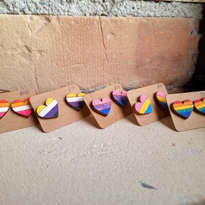Könnte beinhalten: Sechs Paar Ohrringe aus Holz in Herzform, jedes Paar mit einem anderen Design der Pride-Flagge. Die Flaggen sind: lesbisch, bisexuell, nicht-binär, transgender, pansexuell und Regenbogen. Jedes Paar wird auf einer kleinen Karte mit dem Preis von 10 € angezeigt.
