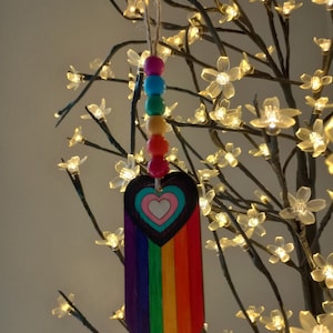 Peut inclure: Une décoration de cœur en bois aux couleurs de l'arc-en-ciel avec un cœur blanc à l'intérieur. La décoration est suspendue à une chaîne de perles en bois colorées.
