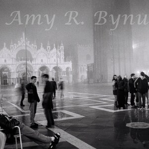 May include: A black and white photograph of a group of people standing in a large, open square in front of a building with arches. The square is paved with large, square tiles. The building is in the background and is partially obscured by fog. The people are all dressed in casual clothing. The photograph is taken at night and the square is lit by streetlights.