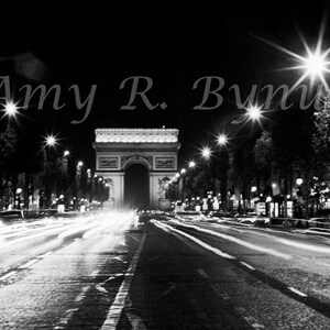 May include: A black and white photograph of the Arc de Triomphe in Paris, France at night. The street is illuminated by streetlights and car headlights.