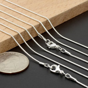 May include: A set of six silver snake chains of varying lengths, displayed on a wooden surface. The chains are arranged in a staggered pattern, with the shortest chain at the top and the longest chain at the bottom. A US quarter is included for scale.