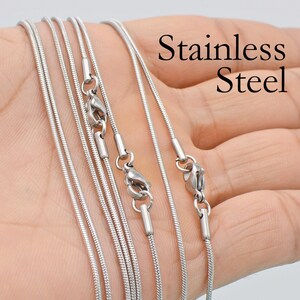 May include: A set of six silver snake chains with lobster clasps. The chains are arranged in a fan shape on a hand. The text "Stainless Steel" is visible in the image.