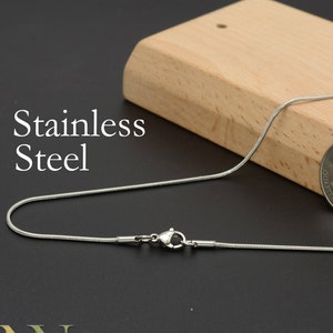 May include: A silver snake chain necklace with a lobster clasp closure. The chain is lying on a wooden surface next to a US dollar coin.