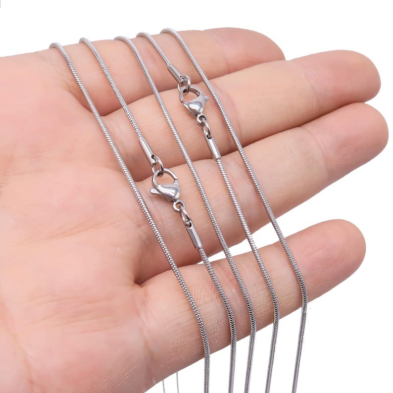May include: A set of four silver snake chains with lobster clasps. The chains are layered on top of each other and are held in a hand.