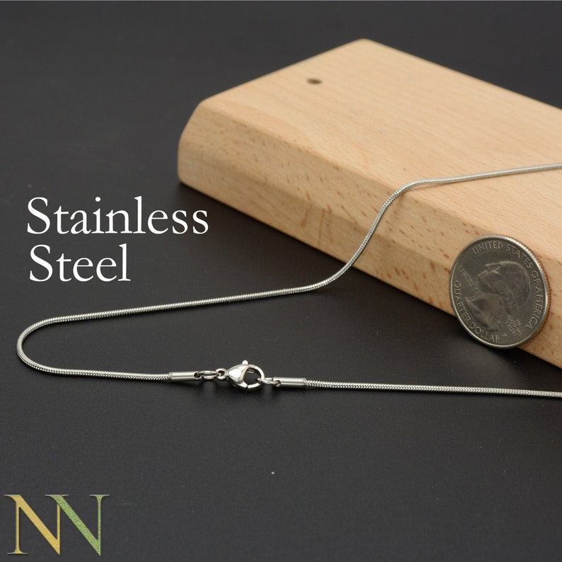 May include: A silver snake chain necklace with a lobster clasp closure. The chain is lying on a wooden surface next to a US dollar coin.