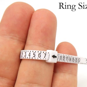 May include: White plastic ring sizer with black numbers from 2 to 17. The sizer is on a finger.