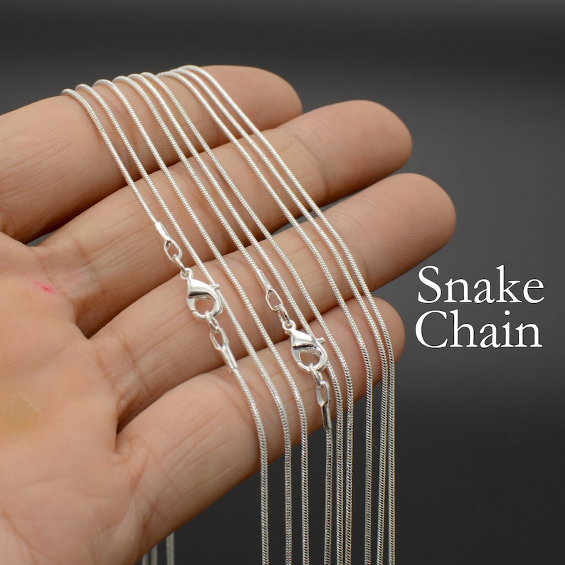 May include: A set of eight silver snake chains, each with a lobster clasp closure. The chains are arranged in a fan shape on a hand, with the clasps at the bottom. The chains are all the same width, but vary in length.  Snake Chain.
