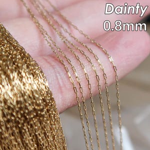 Puede incluir: Se muestran varias hebras de cadena de color dorado, cada una con una anchura de 0,8 mm, en una mano. La cadena está hecha de pequeños cuadrados unidos. El texto "Dainty 0.8mm" es visible en la imagen.