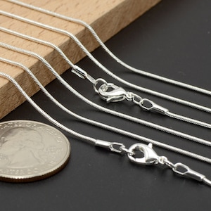 May include: A set of six silver snake chains of varying lengths, displayed on a wooden surface. The chains are made of a thin, delicate material and feature a lobster clasp closure.