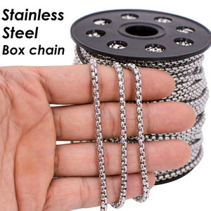 Puede incluir: Un carrete de cadena de caja de acero inoxidable plateado. La cadena se muestra en una mano, y el carrete está en el fondo.