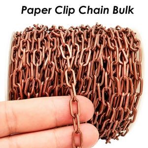 Puede incluir: Primer plano de una cadena de clips de papel de color cobre. La cadena está hecha de clips de papel individuales que están unidos entre sí para formar una cadena continua. La cadena está enrollada y una mano sostiene una sección de la cadena.