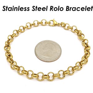 Peut inclure: Un bracelet Rolo en acier inoxydable doré avec un fermoir mousqueton, présenté à côté d'une pièce de monnaie pour comparaison de taille. Le bracelet présente une série de maillons circulaires interconnectés. Le texte "Stainless Steel Rolo Bracelet" est en haut.