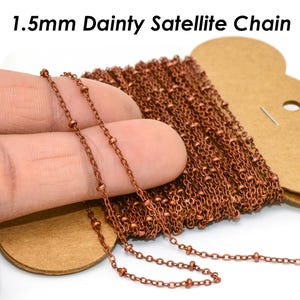 Op de afbeelding: Een spoel van 1,5 mm delicate satellietketting in een koperkleur. De ketting is gemaakt van kleine, ronde kralen die met delicate schakels zijn verbonden. De tekst "1.5mm Dainty Satellite Chain" is zichtbaar bovenaan de afbeelding.