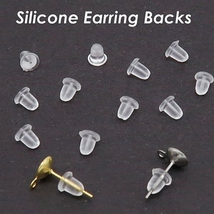 May include: A close-up of a group of clear silicone earring backs. The backs are small and round, with a small hole in the center. There are 13 backs in total, and they are arranged in a random pattern on a black surface.
