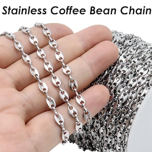 Puede incluir: Primer plano de una cadena de acero inoxidable plateada en forma de grano de café. La cadena está enrollada en un carrete blanco y también se muestra colgada sobre una mano.