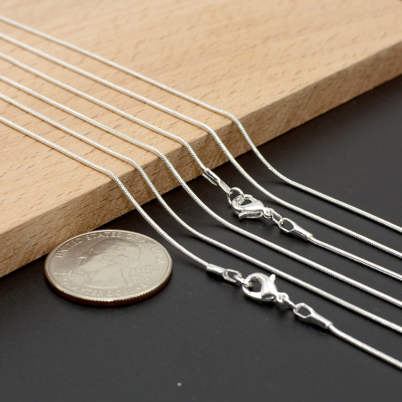May include: A set of six silver snake chains of varying lengths, displayed on a wooden surface. The chains are arranged in a staggered pattern, with the shortest chain at the top and the longest chain at the bottom. A US quarter is included for scale.