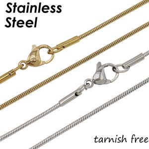 Könnte beinhalten: Nahaufnahme von zwei Schlangenkettchen aus Edelstahl mit Karabinerverschlüssen. Eine Kette ist goldfarben, die andere silberfarben. Der Text "Stainless Steel" und "tarnish free" sind sichtbar.