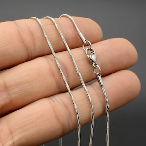 May include: A silver snake chain necklace with a lobster clasp closure. The chain is shown on a hand, with three strands of the chain visible.