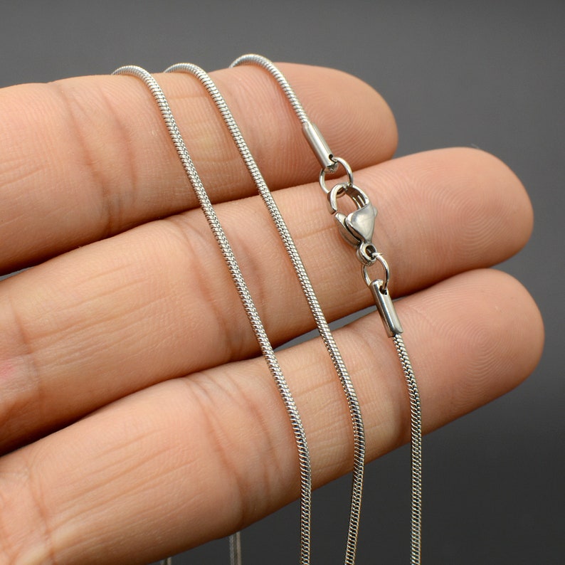 May include: A silver snake chain necklace with a lobster clasp closure. The chain is shown on a hand, with three strands of the chain visible.