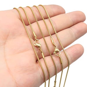 May include: Five gold-colored snake chains of varying lengths, displayed on a hand. The chains are made of stainless steel and have lobster claw clasps.