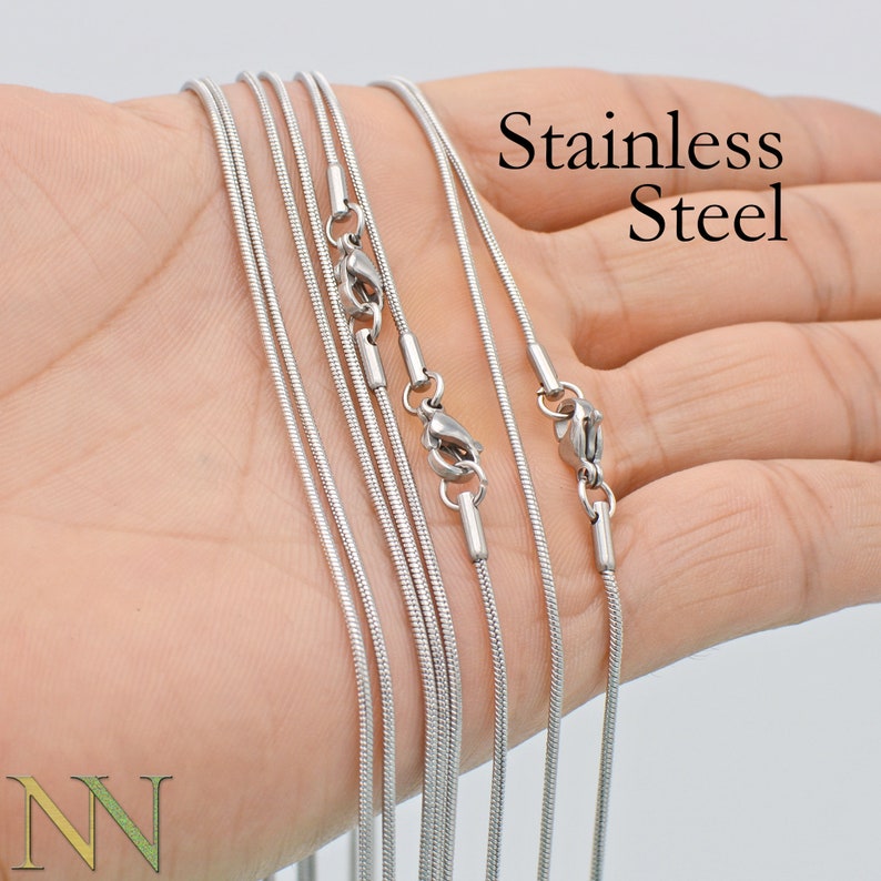 May include: A set of six silver snake chains with lobster clasps. The chains are arranged in a fan shape on a hand. The text "Stainless Steel" is visible in the image.